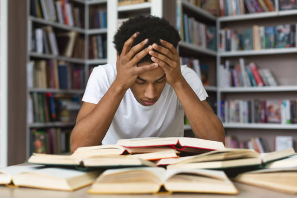 exhausted black guy holding his head and reading book in library